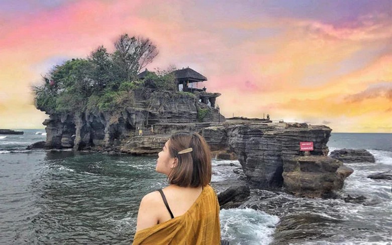 Du khách đứng ở bờ đá ngắm đền Tanah Lot từ xa, nước biển xanh trong, bầu trời đang chuyển sang màu cam