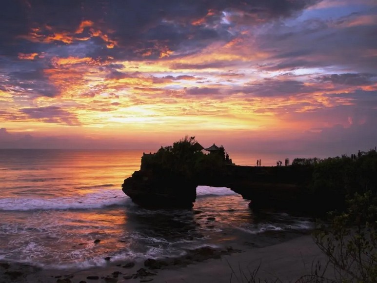 Hoàng hôn từ đền Tanah Lot nhìn ra biển, bầu trời đỏ cam, sóng vỗ chân đền