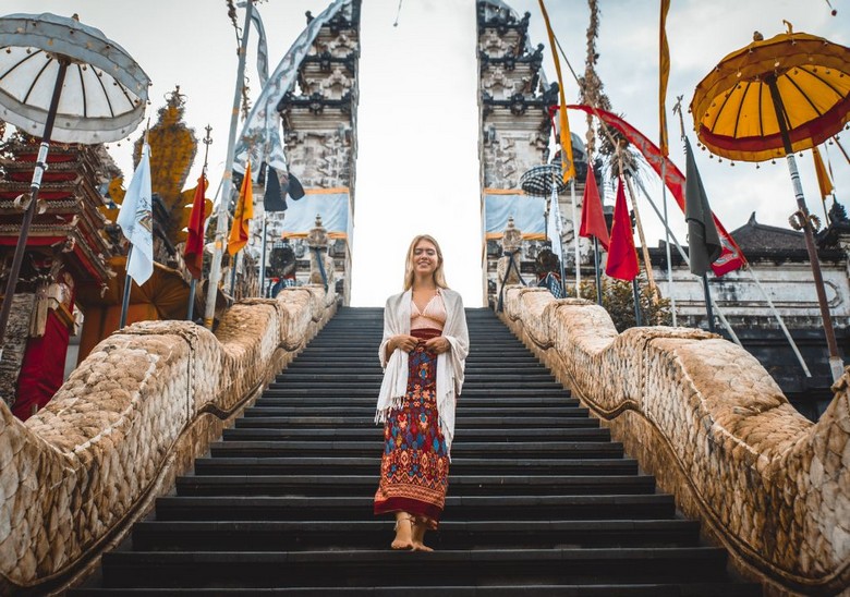 Một du khách mặc váy linen nhẹ đứng trước cổng đền Tanah Lot, có sarong quấn quanh eo