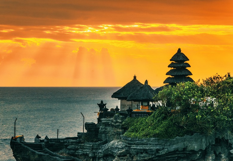 Toàn cảnh đền Tanah Lot nhìn từ xa lúc hoàng hôn, sóng biển vỗ vào chân đá, bầu trời cam đỏ