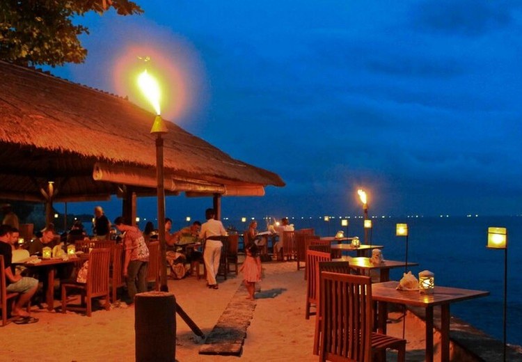 Bãi cát Jimbaran lúc chiều tà, hàng ghế gỗ và bàn ăn dọc mép nước, ánh đèn nến bắt đầu le lói