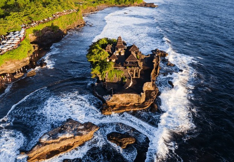 Đền Tanah Lot nhìn từ xa lúc chiều tà, sóng biển vỗ quanh mỏm đá, du khách đứng dọc bờ kè ngắm cảnh