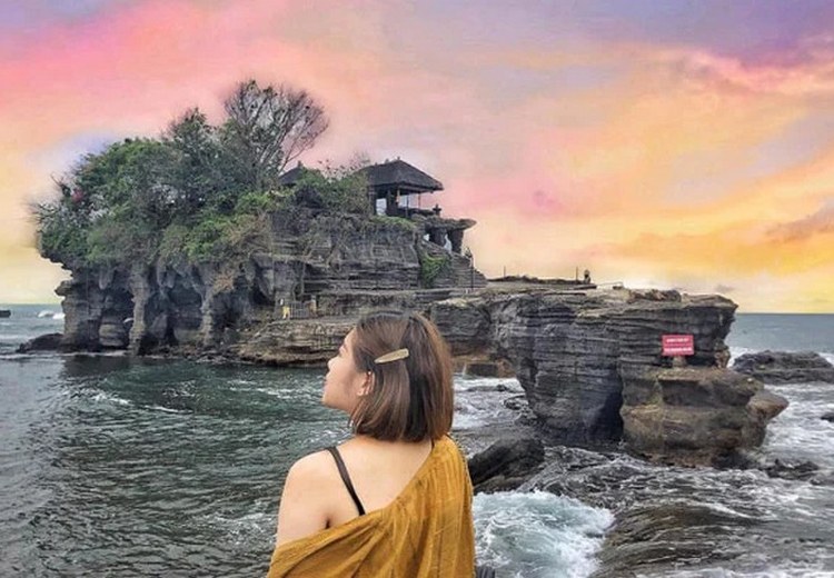 Du khách đứng ở bờ đá ngắm đền Tanah Lot từ xa, nước biển xanh trong, bầu trời đang chuyển sang màu cam