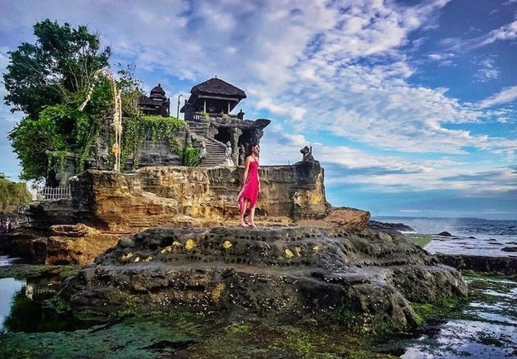 Hoàng hôn Tanah Lot nhìn từ góc thấp, bóng người đứng ngắm phía xa, sóng biển vỗ mạnh vào tảng đá