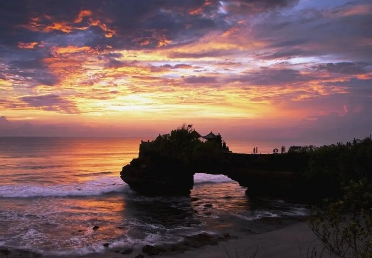 Hoàng hôn từ đền Tanah Lot nhìn ra biển, bầu trời đỏ cam, sóng vỗ chân đền