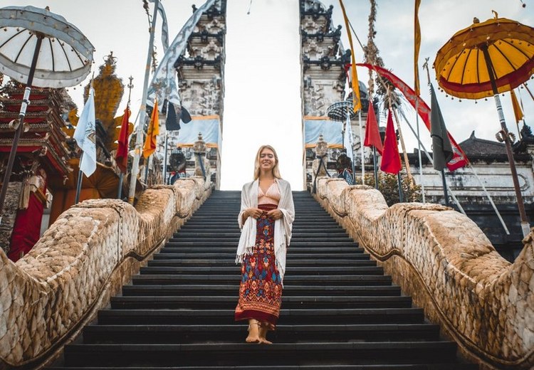 Một du khách mặc váy linen nhẹ đứng trước cổng đền Tanah Lot, có sarong quấn quanh eo