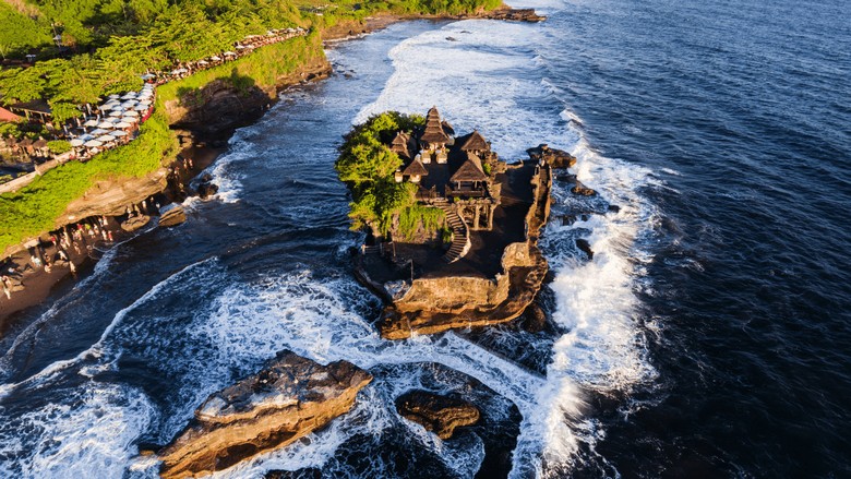 Đền Tanah Lot nhìn từ xa lúc chiều tà, sóng biển vỗ quanh mỏm đá, du khách đứng dọc bờ kè ngắm cảnh